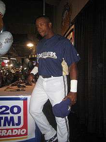 A man wearing a navy blue Brewers jersey and white pants holds a navy blue cap in his hand as he leaves a table after completing a radio interview.