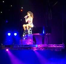 Rihanna is standing on a grand piano on a stage illuminated with purple light.