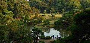 A panoramic view of the Rikugi-en Garden from the Fujishiro-toge hill vantage point. Green trees surround a serene lake.