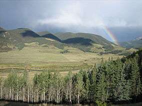A small river winds through mountains under a rainbow.