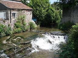 Water flowing through a channel and over a weir between a building and a wall. Vegeatation on both sides of the water.