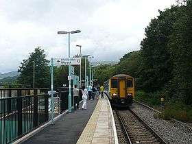 View along country station platform with bilingual exit signage. Passengers wait to board a two carriage train, which is stationary on the single track. Platform, train and track curve slightly right to left. Trees in full leaf stand alongside the track, with mountains in the distance.