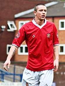 Rory Patterson in red F.C. United home kit playing on pitch, in front of brick buildings.