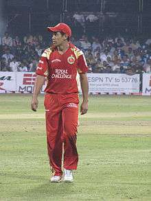 An cricketer wearing red outfit and shirt. Spectators can be seen in background.