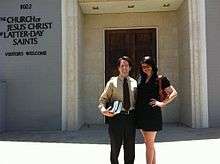 Man with light skin and dark hair and woman with light hair and light skin stand in front of a building with a sign that reads "The Church of Jesus Christ of Latter-day Saints; Visitors Welcome"