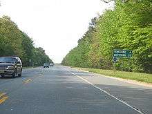 A two lane road running through woodland. A green sign along the road reads Vineland 6 Deptford 35