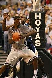A African-American teenage basketball player wearing a gray uniform looks over his shoulder at another playing in a blue uniform. Behind them are fans and a basketball hoop with the word "HOYAS" on it.
