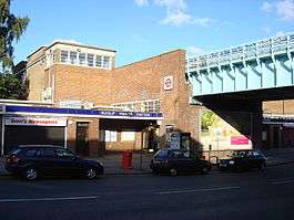 A brown-bricked building with a rectangular, dark blue sign reading "RUISLIP MANOR STATION" in white letters all under a light blue sky