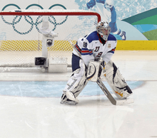 An ice hockey goaltender is on the ice in front of his net. He is wearing a white sweater with the letters "USA" on the front in blue.