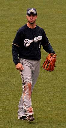 A man wearing a navy blue Brewers jersey, gray pants, navy blue cap, and outfielder's glove on his left hand walking in the outfield.