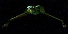 Forward shot of a starship in the black of space with distant stars in the background. The ship is primarily one color, save for a decorative pattern on its underside. The center portion of the ship is bulbous, with thin, down-stretched wings capped by long guns.