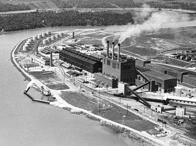 A factory with three smoking chimneys on a river bend, viewed from above