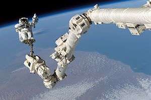 A photograph of a long, white mechanical arm stretching out above a mostly blue planet displaying white clouds and brown terrain all under a black expanse