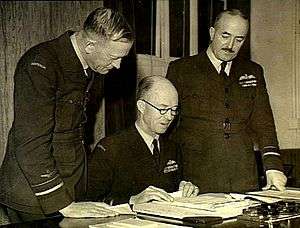 Thee men in military uniforms, two standing and one seated, looking at papers on a desk