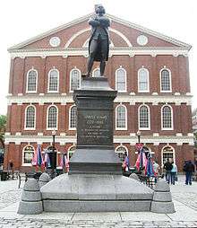 A statue on a pedestal of a man standing with his arms crossed. An inscription on the pedestal reads, "Samuel Adams, 1722–1803. A Patriot. He organized the Revolution and signed the Declaration of Independence." Behind the statue is a three story brick building with many windows.