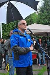 A man behind a microphone stand, wearing glasses and a blue jacket; behind him is a man holding up an umbrella