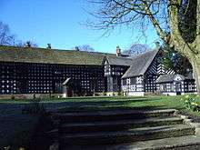 A two-storey black and white timbered building in an L-shape. A wide set of stone steps in the foreground leads onto a grassed area in front of the house.