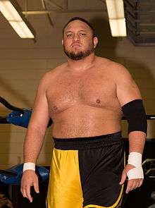 An adult Samoan male posing while wearing orange and black wrestling trunks.