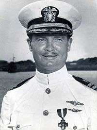 Head and shoulders of a white man with a thin mustache wearing a white peaked cap, its black visor decorated with oak leaves, and a white military jacket with dark shoulder boards and one medal and two winged pins on the left breast.