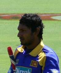 Portrait picture of a dark skinned man wearing blue and yellow Sri Lankan cricket kit, with a partially visible cricket bat under his arm.