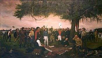A group of men are gathered under a large tree.  One man lays on the ground under the trees, with his bare foot exposed.