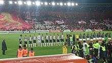 Two football teams and a refereeing team lining up in a football stadium pitch. The leftmost team wears black and white striped shirts, black shorts and white socks, while the other wears white shorts, red socks and gray jackets. The refereeing trio wears yellow shirts and black socks. People with cameras can be seen in the foreground. A stand below lit floodlights is seen in the background, packed with people in red clothes holding red and white banners and paper pieces.