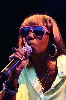 Woman singing into a microphone wearing bright pink sunglasses, large earrings, two small necklaces, and a pale green shirt.
