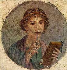 Round painting of a woman with curly hair wearing a gold hairnet while holding a wax writing tablet. She has the stylus in her right hand and the tip in her mouth.