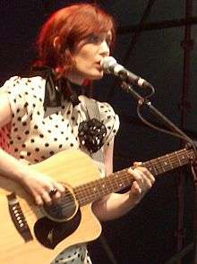 29-year-old Blasko is shown in an upper body shot. She is leaning and singing into a microphone on its stand to her left while looking forward. She is holding her guitar with her right hand plucking the strings and her left low on the fret board. She wears a cream-coloured dress with dark polka dots. She wears a dark, twisted ring on her right middle finger.