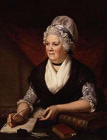 Half-length portrait of an elderly woman sitting at a desk surrounded by books and papers and holding a quill.