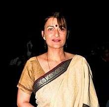 A middle aged Indian woman with sharp features and grey colored eyes, wearing a yellow sari. Her hair is bound behind her.