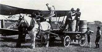 A man refuels a single-engined biplane in a field using barrels on a horse-drawn cart