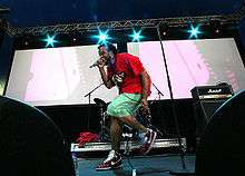 A man wearing a red T-shirt and light green shorts on a stage singing into a wired microphone. A white screen and blue stage lights serve as his backdrop in an evening setting.
