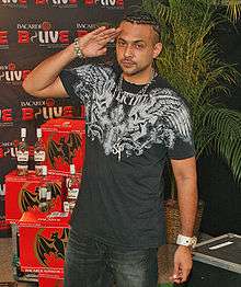 A man saluting with his right hand and wearing a black graphic T-shirt and black jeans. In the background is a palm tree, a few bottles of rum, and a screen with branding across it.