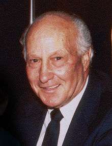 A balding older man, smiling, wearing a suit jacket and tie