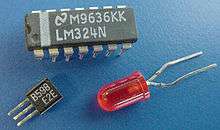A small square plastic piece with three parallel wire protrusions on one side; a larger rectangular plastic chip with multiple plastic and metal pin-like legs; and a small red light globe with two long wires coming out of its base.