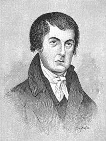 A man with short, black hair wearing a black jacket and vest and white shirt and tie