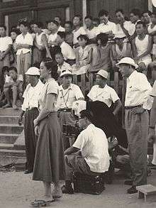 A crowd of people gathered at a film location shoot: in the background, slightly out of focus are many adults and children, some standing, some sitting on stone steps; in the left foreground is a young Japanese woman, Hara, in a white blouse and dark dress, with camera crew behind her; a middle-aged Japanese man, Ozu, in dark pants, white shirt and floppy hat stands at far right foreground.