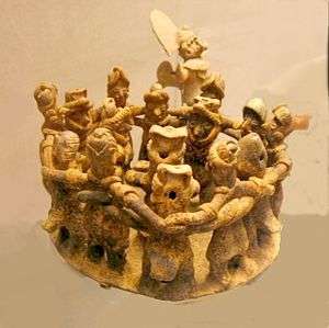 A ring of twelve dancing figures, arms interlocked around each other's shoulders. They surround one musician in the centre of the ring, and a second musician stands behind them.