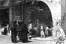 From left to right: A boy staring out from a store's window sill beneath which a lamb is walking by; three fully veiled women are conversing on the street; beneath an olive grove jutting out of a large stone archway and beside a fountain, a man is walking, a woman is collecting water from the fountain, and two young boys are standing and smiling; a young girl walking on the street