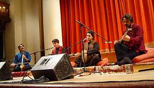 The four original ensemble members sit atop rugs and cushions onstage. The men all have their respective instruments, with microphones and monitor speakers before them. Behind them is a large, orange drapery.