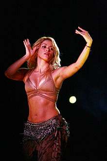 A woman with golden hair is striking a pose and waving her hands in the manner of a belly dancer. She is dressed in a flesh-toned bra and a brown fringe-adorned skirt.