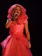 A lady is singing a song dressed in a hooded fuchsia gown. She has a mic in her hand.