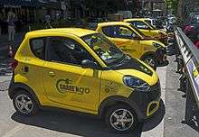 A line of small two-door yellow cars parked diagonally against a curb. Several have curled red charging cords connecting them with a charging station on the curb. On the side of the one nearest the camera is written the words "Share N' Go: the new mobility" with a stylized electric cord curling around the words on their left