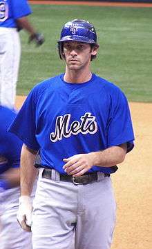 A man in a blue baseball jersey with "METS" on the chest, blue batting helmet, and light-colored pants with a glove on his right hand.