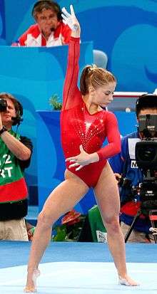 A female gymnast in a red leotard, looking down and to her left, with her right hand raised and her left hand across her waist.  In the background sit a number of people including a cameraman, a photographer, a commentator and a judge