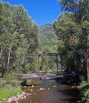 A short dark metallic bridge with rectilinear and diagonal elements over a stream with woods on either side and a wooded mountain in the background