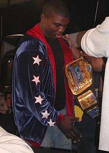 An African American man is facing to the right and holding up a professional wrestling tag team championship, engraved with the words "World Wrestling Entertainment Tag Team Champions". He is wearing light blue jeans, a black T-shirt, and a dark blue jacket, with a red lining, and 5 white stars, outlined in red, are visible upon the left sleeve.