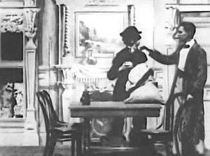 A frame of the black-and-white film. Sherlock Holmes enters his parlour and taps the shoulder of a burglar who is collecting Holmes' tablewares into a sack. Holmes is wearing a dressing gown and smoking a cigar, the thief is dressed in black.