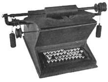 A wooden typewriter with a "QWE-TY" keyboard composed of circular, white keys. The platen rests on top of the device, connected to a bar which extends several inches beyond the sides of the machine. Counterweights are suspended from each end of the bar, to which several wheels and gears are also attached.